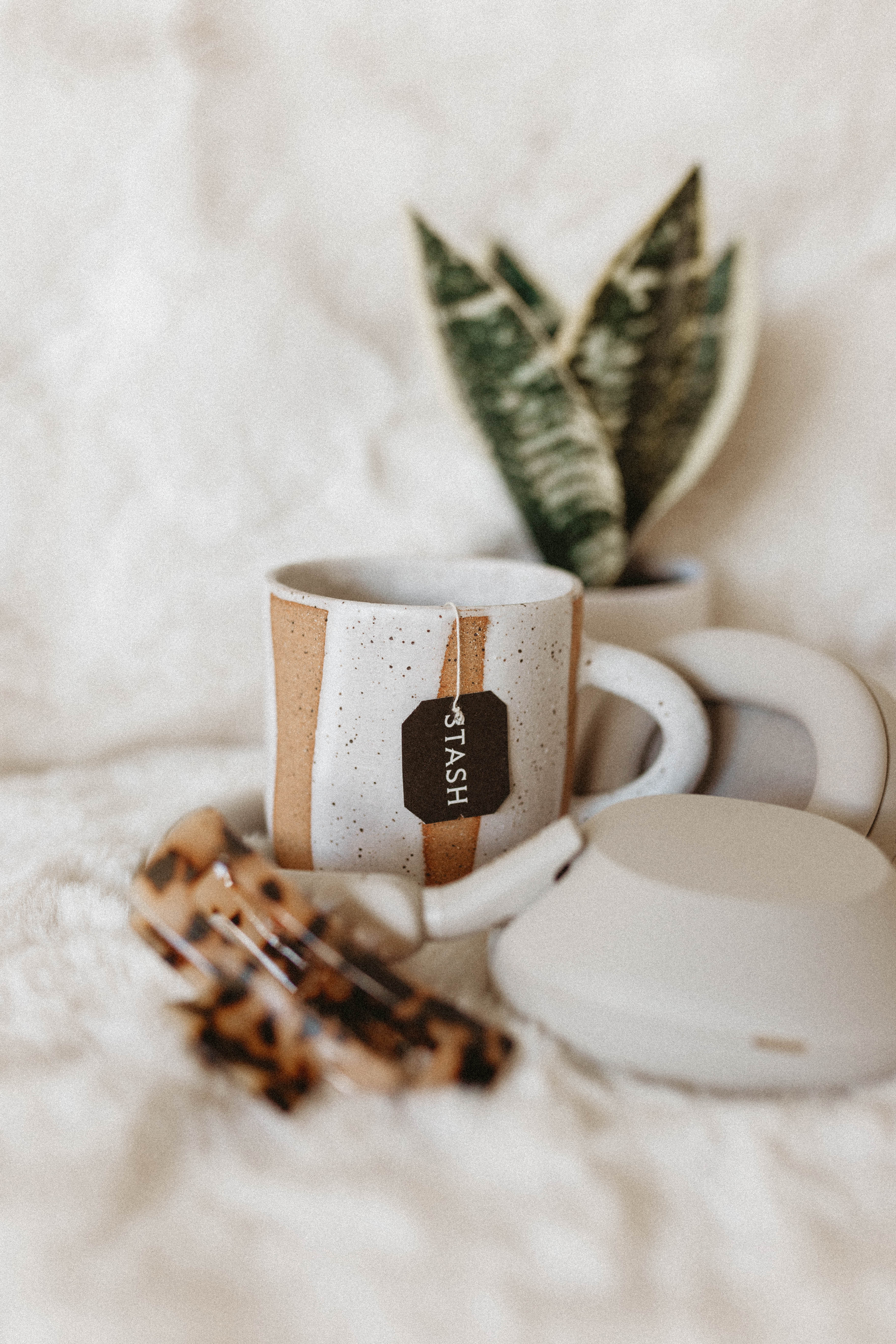 Ceramic mug with 'TASH' branding, teapot, and plant on a soft surface