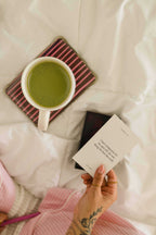 A hand with a floral tattoo holds a small white card with printed text over a bed of white sheets. A white mug filled with green matcha tea rests on a pink-and-white striped coaster nearby, with a black phone and pink knit fabric in the scene.