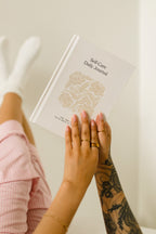 A person holds a white hardcover journal titled 'Self Care Daily Journal' with a beige floral illustration. The photo includes the person's hands, tattooed forearm, pink striped pajama pants, white socks, and bare feet against a soft cream background.