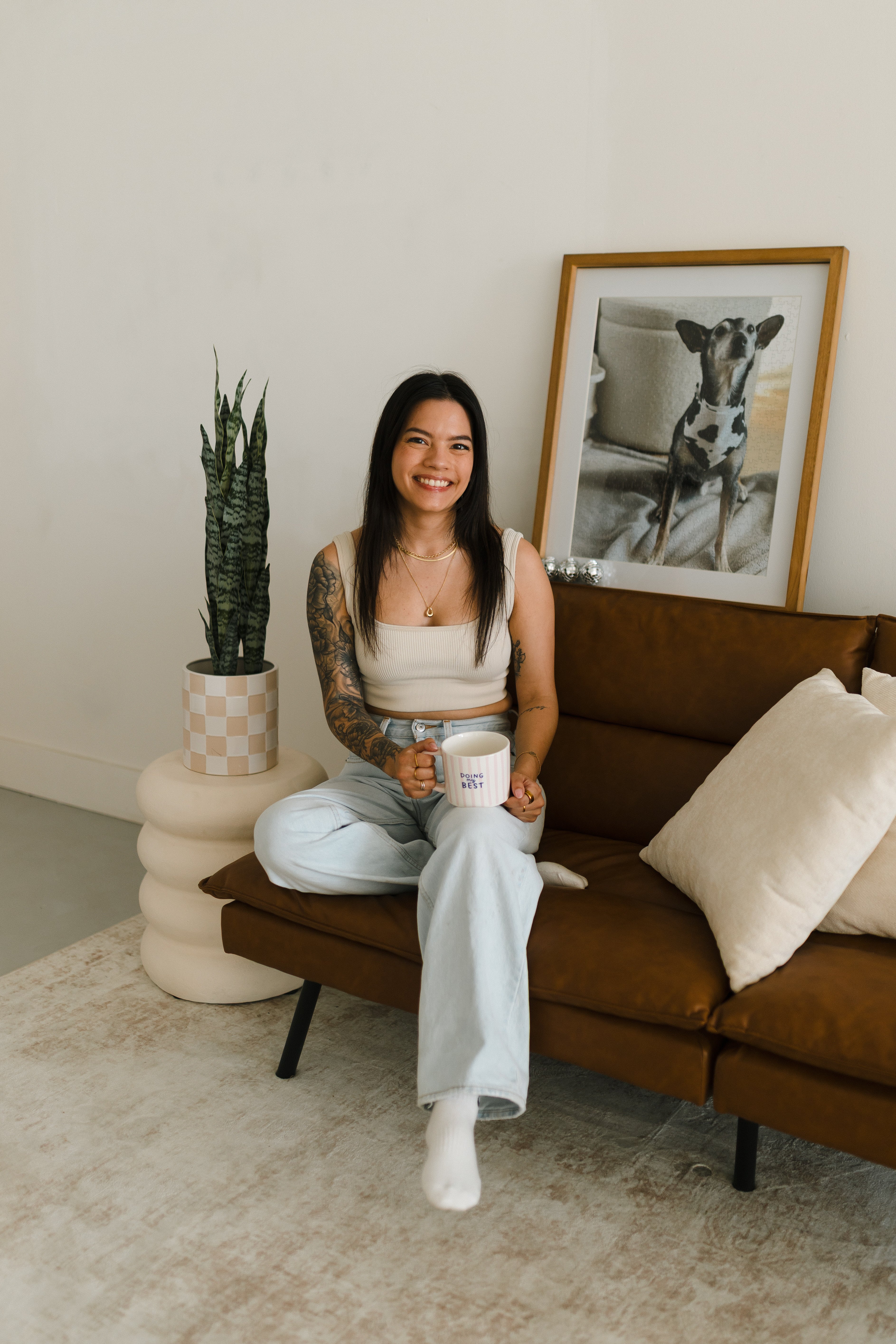 A woman with tattoos sits on a brown leather sofa in a modern living room setting, holding a white mug with blue text saying "doing my best". She is wearing a beige crop top and light blue jeans.