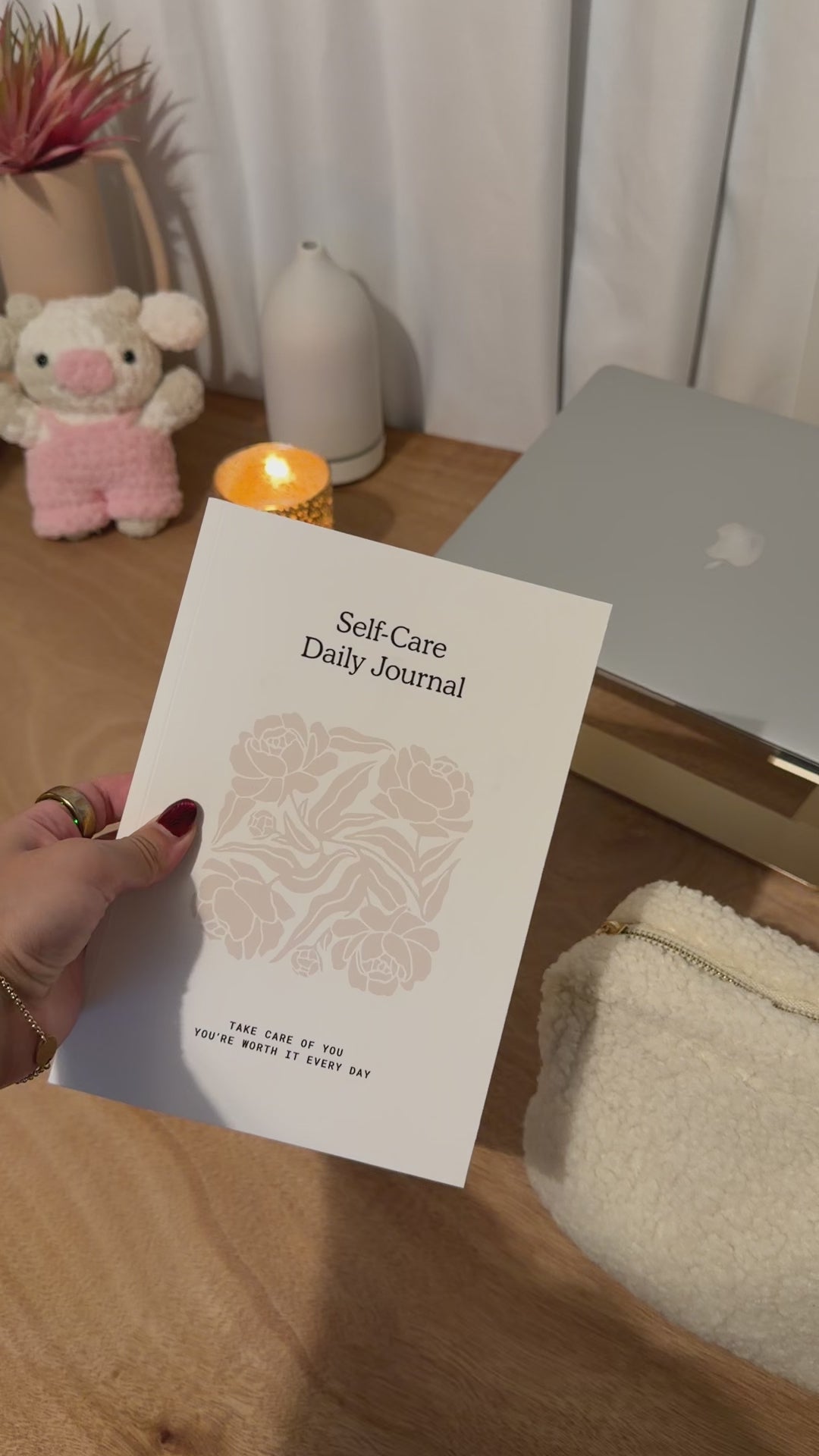 Hand holding a white Self-Care Daily Journal, featuring floral design and motivational quote, on a wooden desk.